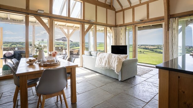Il s'agit d'une pièce spacieuse, dotée d'un plafond haut et de grandes fenêtres qui offrent une vue sur la campagne. On y trouve un coin repas avec une table et des chaises, ainsi qu'un coin salon avec un canapé et un téléviseur.