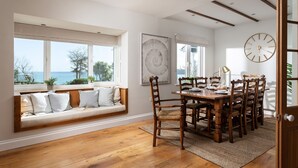 A dining area with a wooden table and chairs, a large wall clock, and a view of the sea through large windows. - Point House - sleeps 8 guests  in 4 bedrooms (Charlestown,)