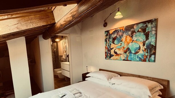 A bedroom with a wooden ceiling, a bed with white linens, a bathroom with a mirror and towel rack, and a colorful abstract painting on the wall.