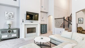 Ein großzügiges Wohnzimmer mit Kamin, einem darüber angebrachten Fernseher, einer Treppe zu einer oberen Ebene und einer gemütlichen Sitzecke mit einem weißen Sofa und einem runden Couchtisch.