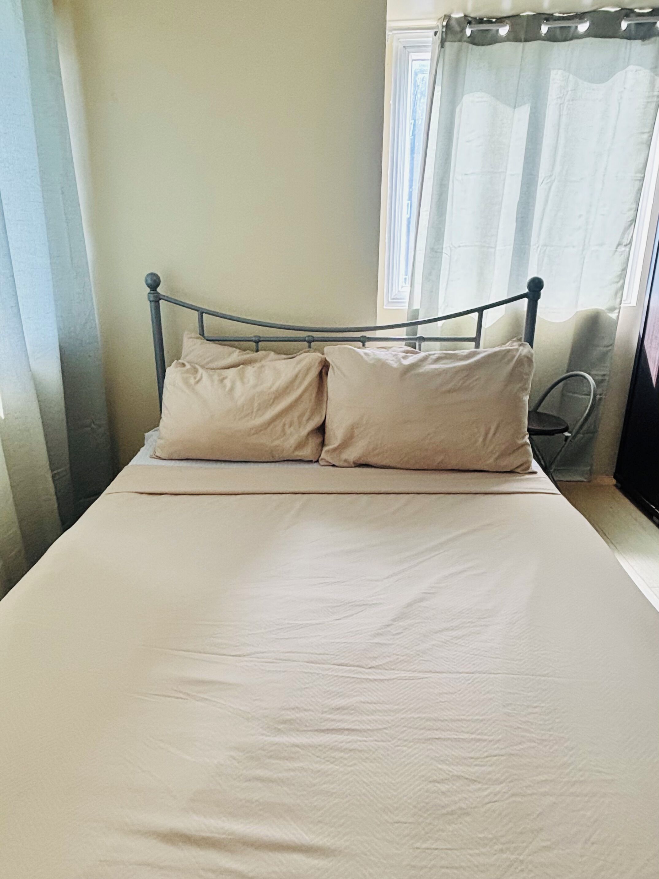 Un lit avec un cadre en métal et deux oreillers, une chaise et une fenêtre avec des rideaux.