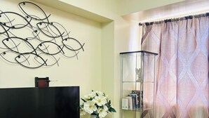 A living room with a flat-screen TV on a black stand, a white vase with flowers, a glass cabinet, and a patterned curtain. - Charming Iloilo City Condo for Rent - Entire House (Iloilo)