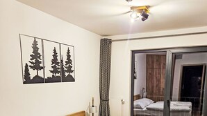 Een net opgemaakt bed met witte lakens, een houten hoofdbord en een nachtkastje met een lamp. Aan de muur hangt een lijst met een foto van bomen, en een schuifdeur leidt naar een badkamer.