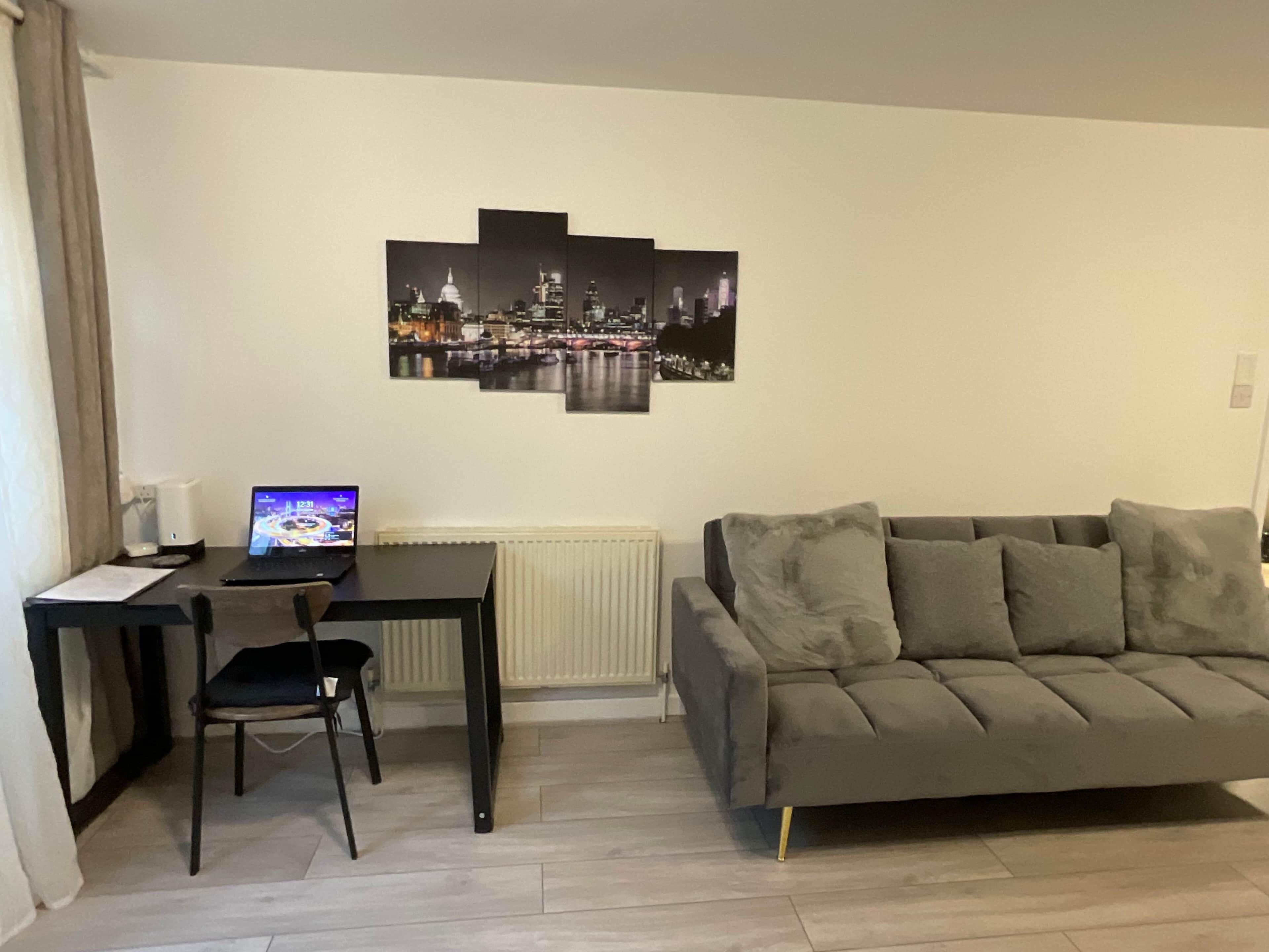 A living room with a grey sofa, a black desk with a laptop, a chair, and a cityscape artwork on the wall.