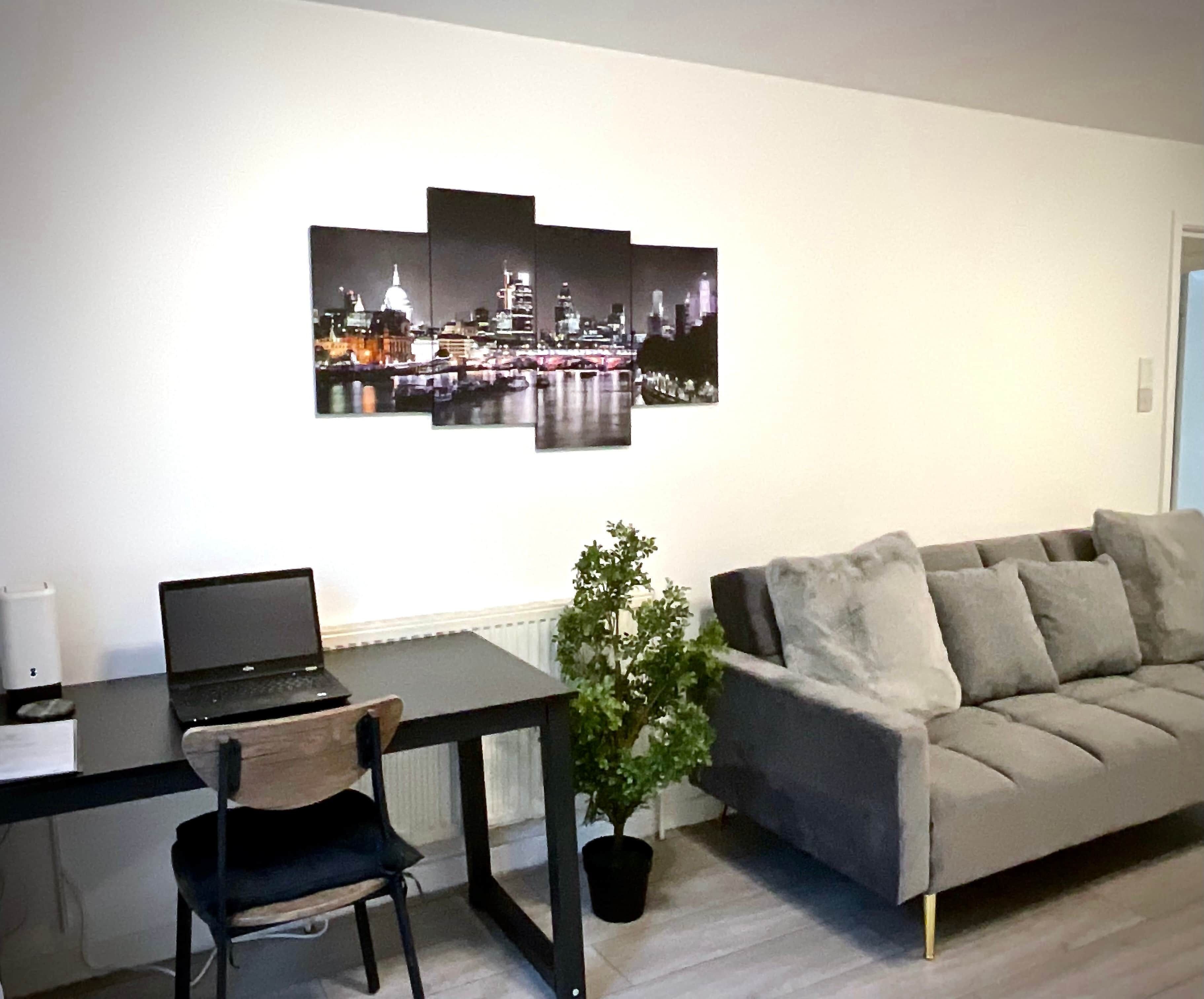 A living room with a grey sofa, a black desk with a laptop, a small potted plant, and a cityscape artwork on the wall.