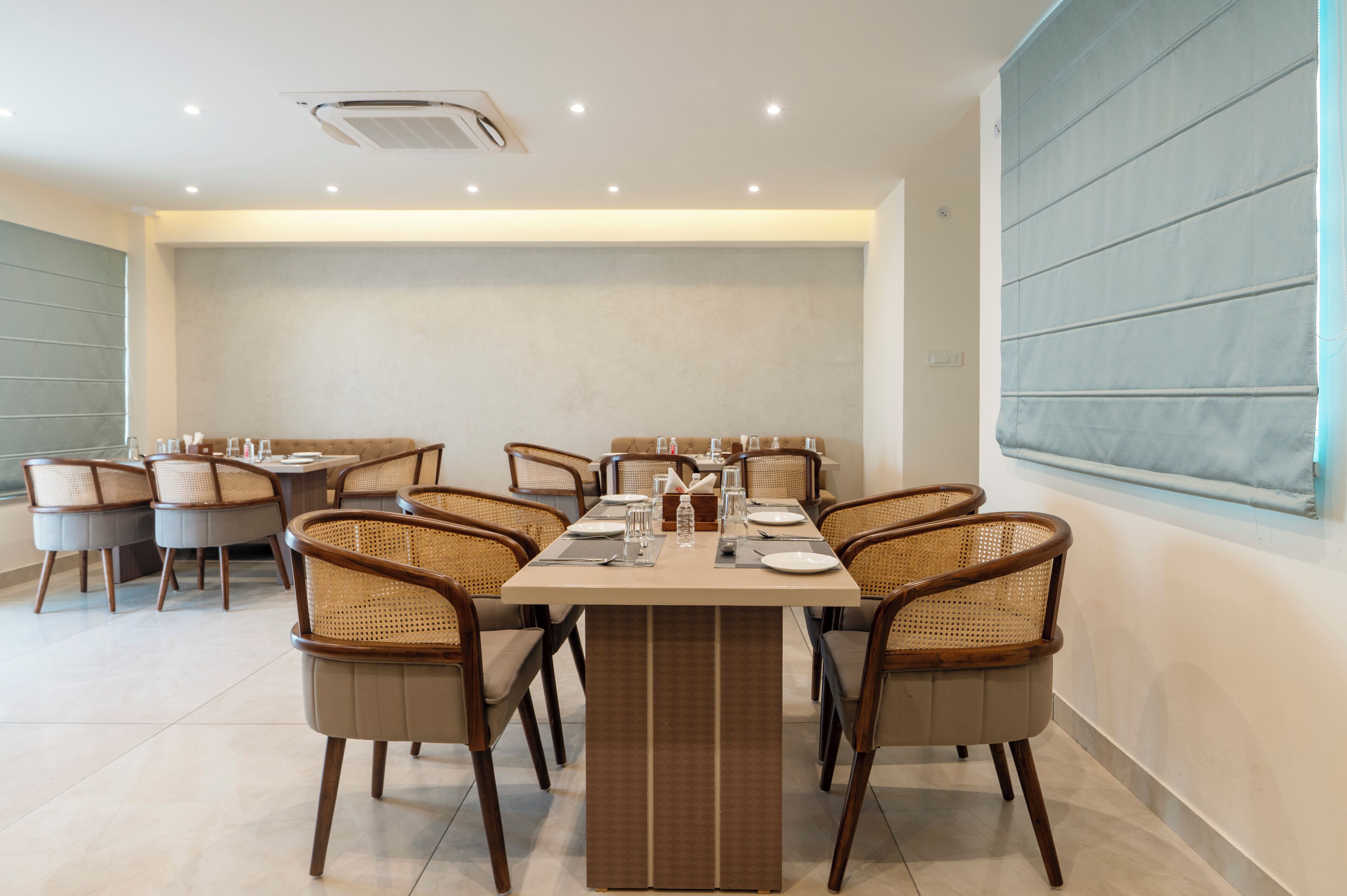 A dining area with a rectangular table set for six, surrounded by wicker chairs, with a large window on the right and a plain wall on the left.