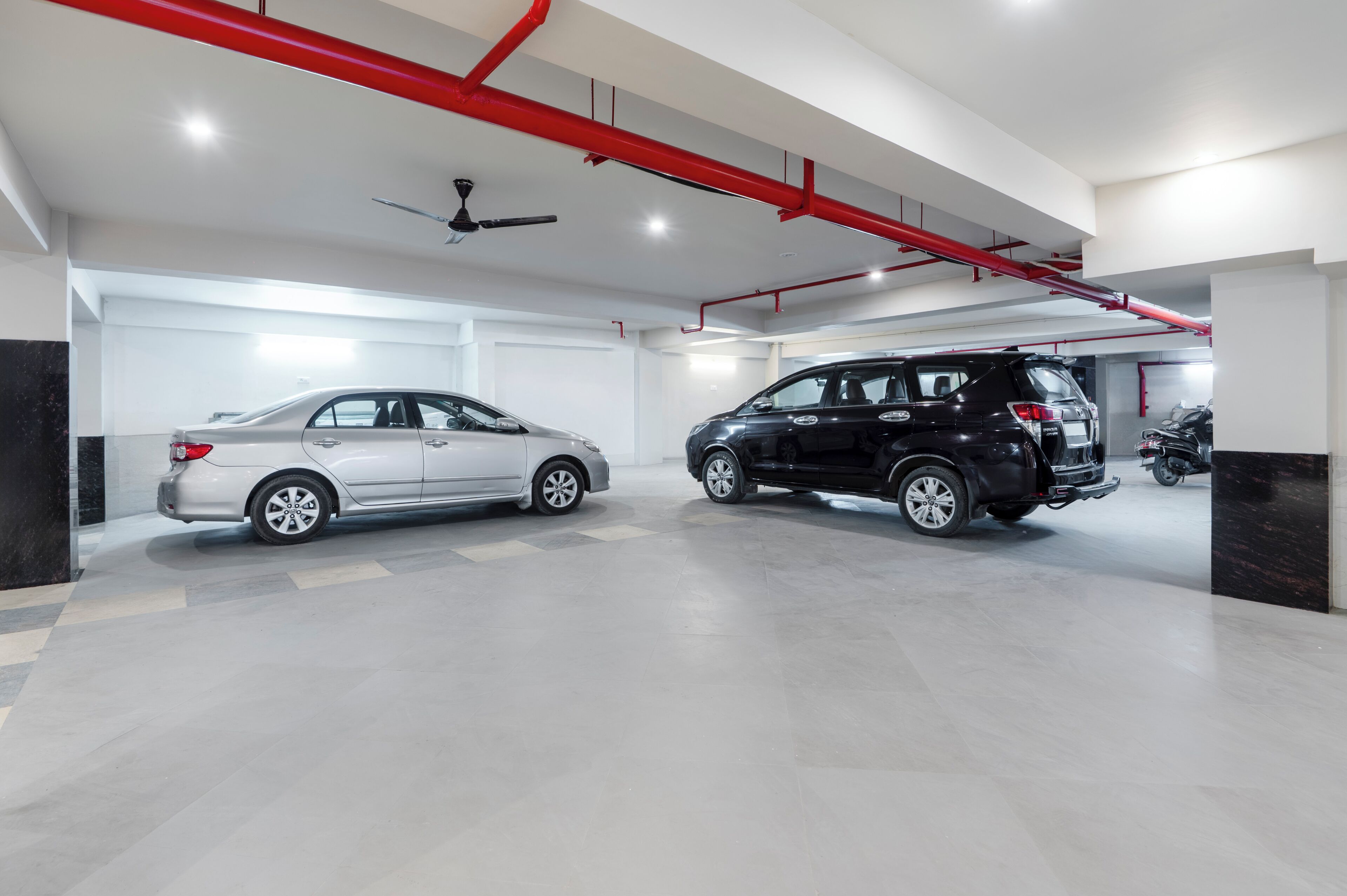 A modern garage with two cars parked, a ceiling fan, and red pipes.