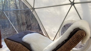 Una habitación con techo de vidrio geométrico, una silla tejida con cojín blanco y vistas a un área boscosa.