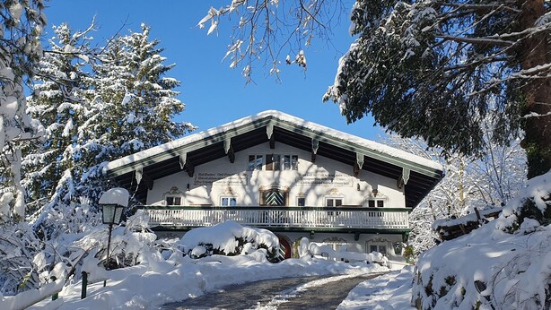 A traditional chalet with a balcony, surrounded by snow-covered trees and a clear sky.