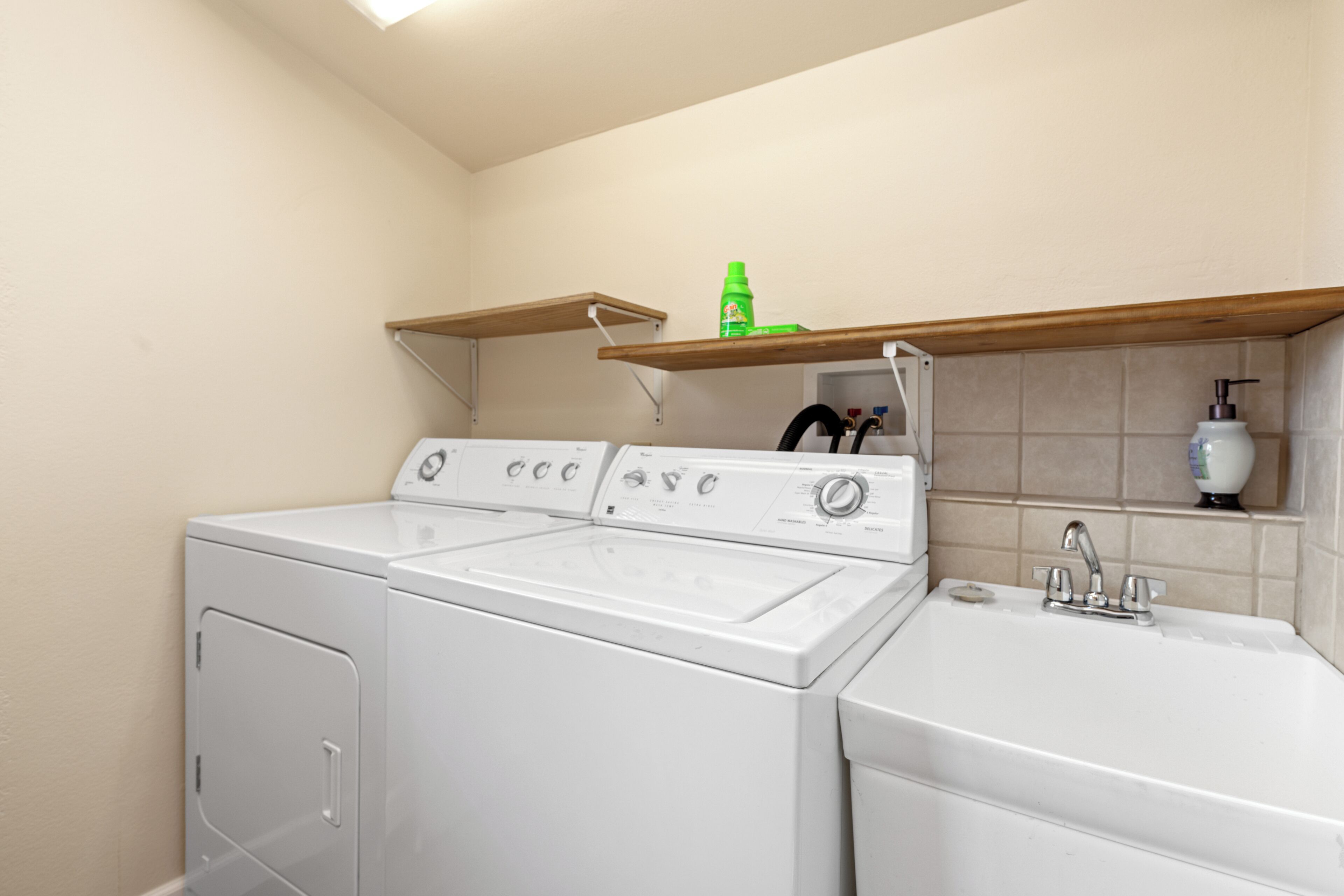 A laundry area with a front-loading washer and dryer, a sink, and a shelf above the dryer holding a green bottle and other items.
