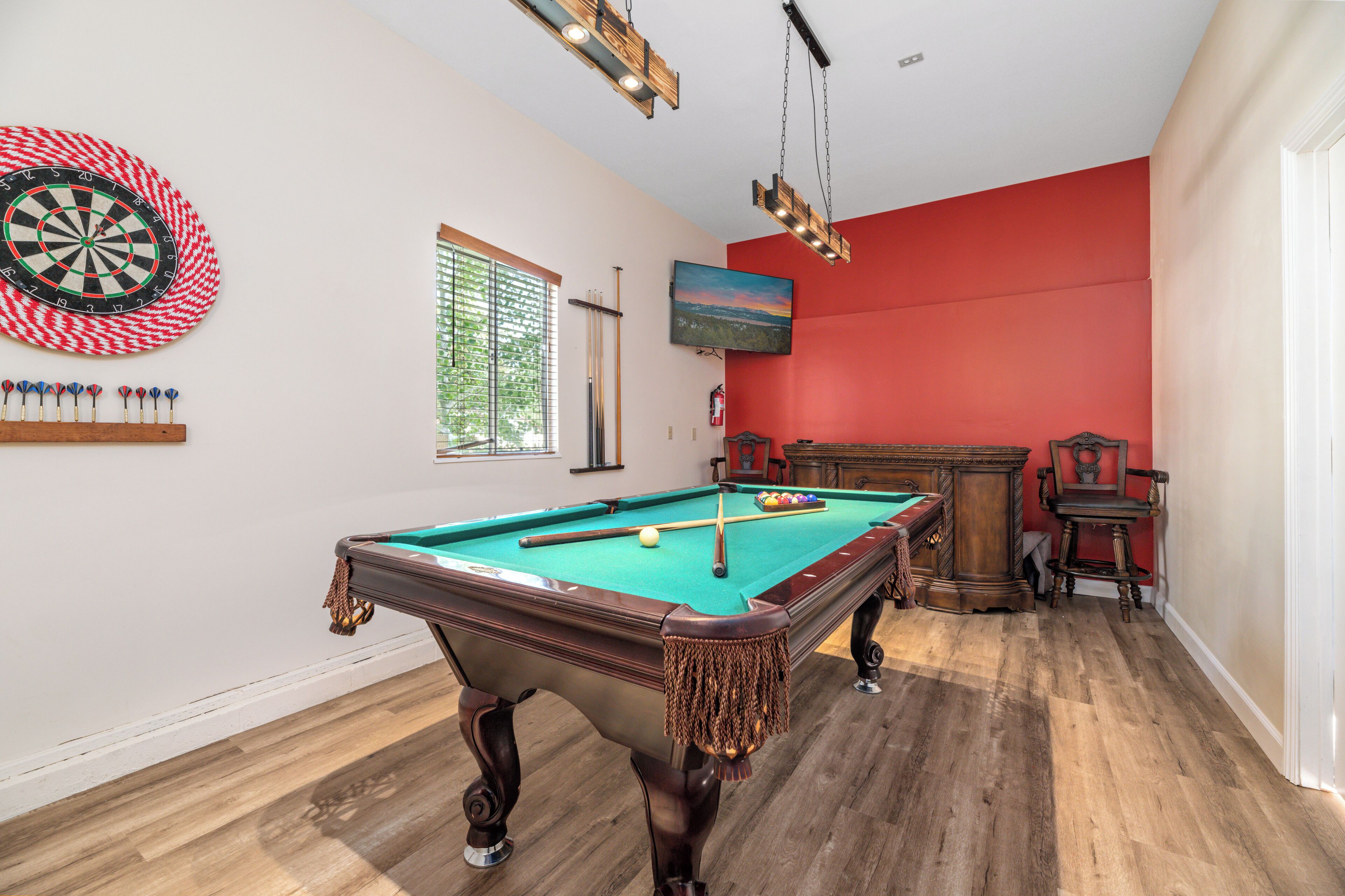 A pool table with a dartboard and pool cues, a wooden floor, and a red wall.