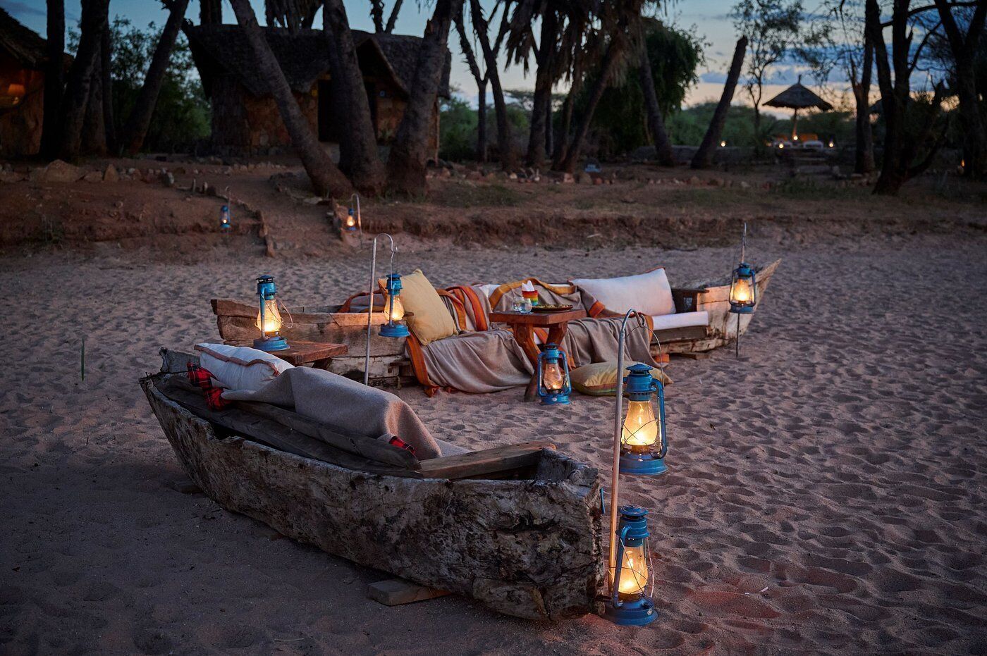 A beach setting with a rustic wooden boat, lounge chairs, and lanterns providing ambient lighting.
