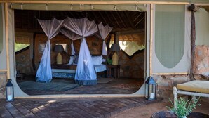 A rustic bedroom with a stone wall, a canopy bed, a nightstand with a lamp, and a lantern on the floor. - Saruni Rhino (Kauro)