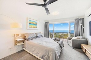 A modern bedroom with a bed, a desk, a chair, and a balcony with a view. - Wake to the Waves - Oceanfront Apartment (Fraser Coast)