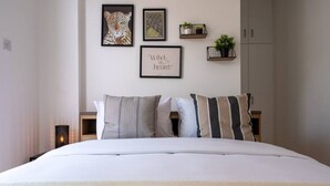 A neatly made bed with a white comforter and striped pillows, a framed artwork of a leopard, a framed quote, and small potted plants on wall-mounted shelves. - Chloris 3- Bedroom Apartment in Larnaca (Larnaca)