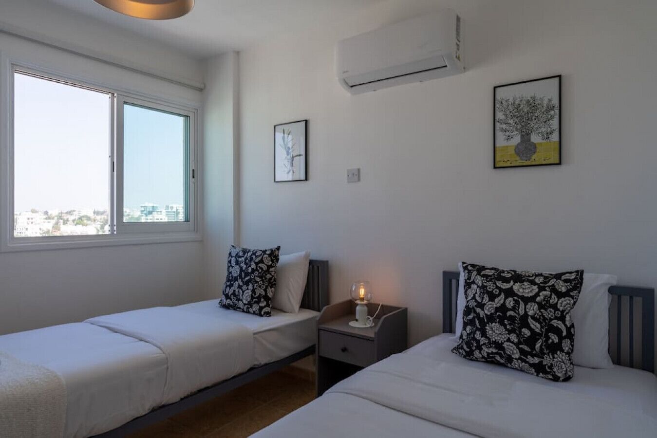 A bedroom with two beds, a nightstand with a lamp, an air conditioner, and a window with a city view.