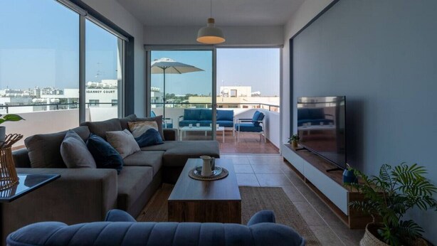 A modern living room with a sofa, coffee table, and TV. There are large windows and a balcony with outdoor seating. - Chloris 3- Bedroom Apartment in Larnaca (Larnaca)