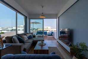 Ein modernes Wohnzimmer mit einer Couch, einem Couchtisch und einem Fernseher. Große Fenster und ein Balkon mit Sitzgelegenheit sind vorhanden.