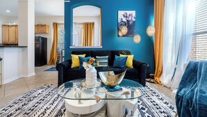 Een moderne woonkamer met een blauwe accentmuur, een zwarte bank met gele kussens, een glazen salontafel en een keuken op de achtergrond.