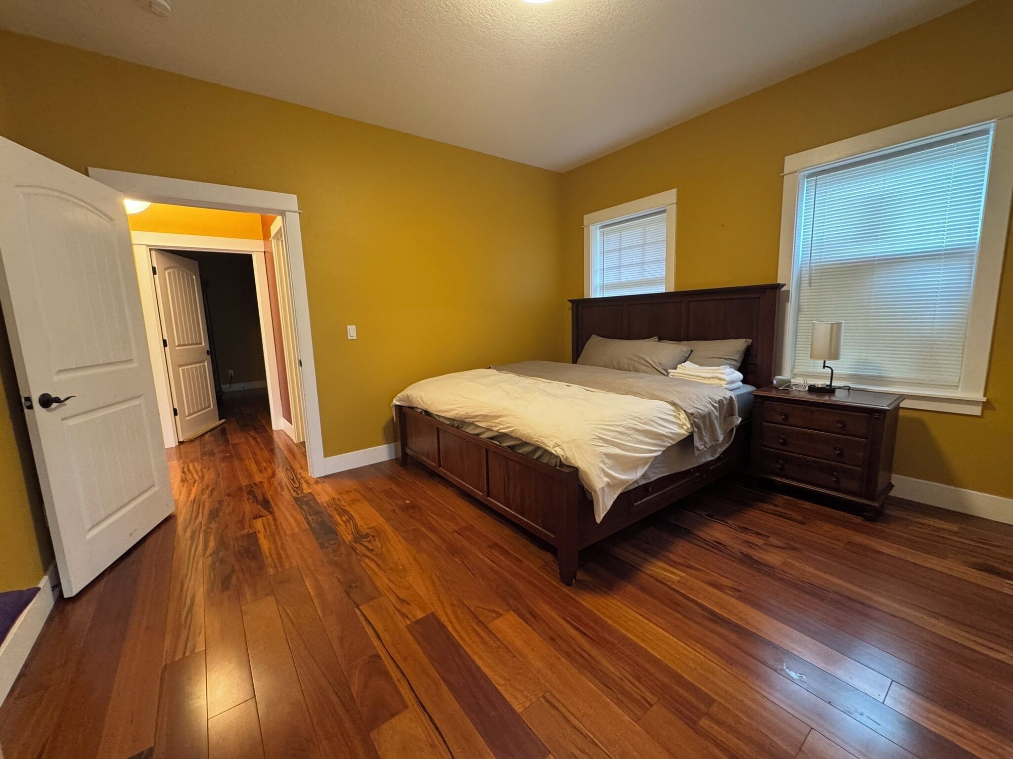 A bedroom with a wooden floor, a bed with a headboard, a dresser, and two windows with blinds.