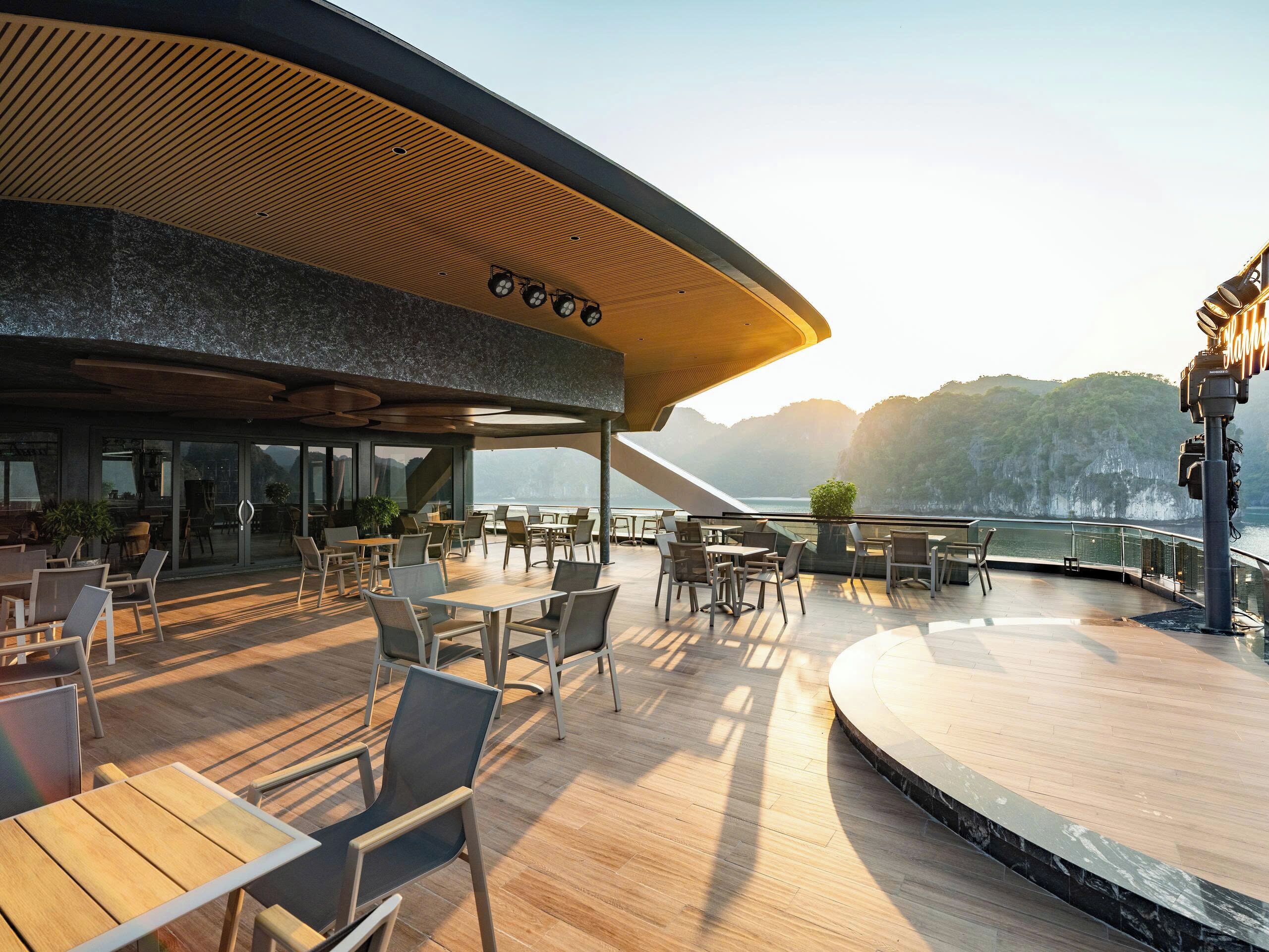 An outdoor dining area with wooden tables and chairs, overlooking a body of water and hills.