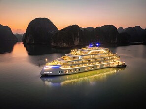 A large, multi-deck cruise ship with illuminated decks and a prominent bow, sailing in calm waters surrounded by rocky cliffs. - Tulip Cruise by HAG (Tuan Chau)