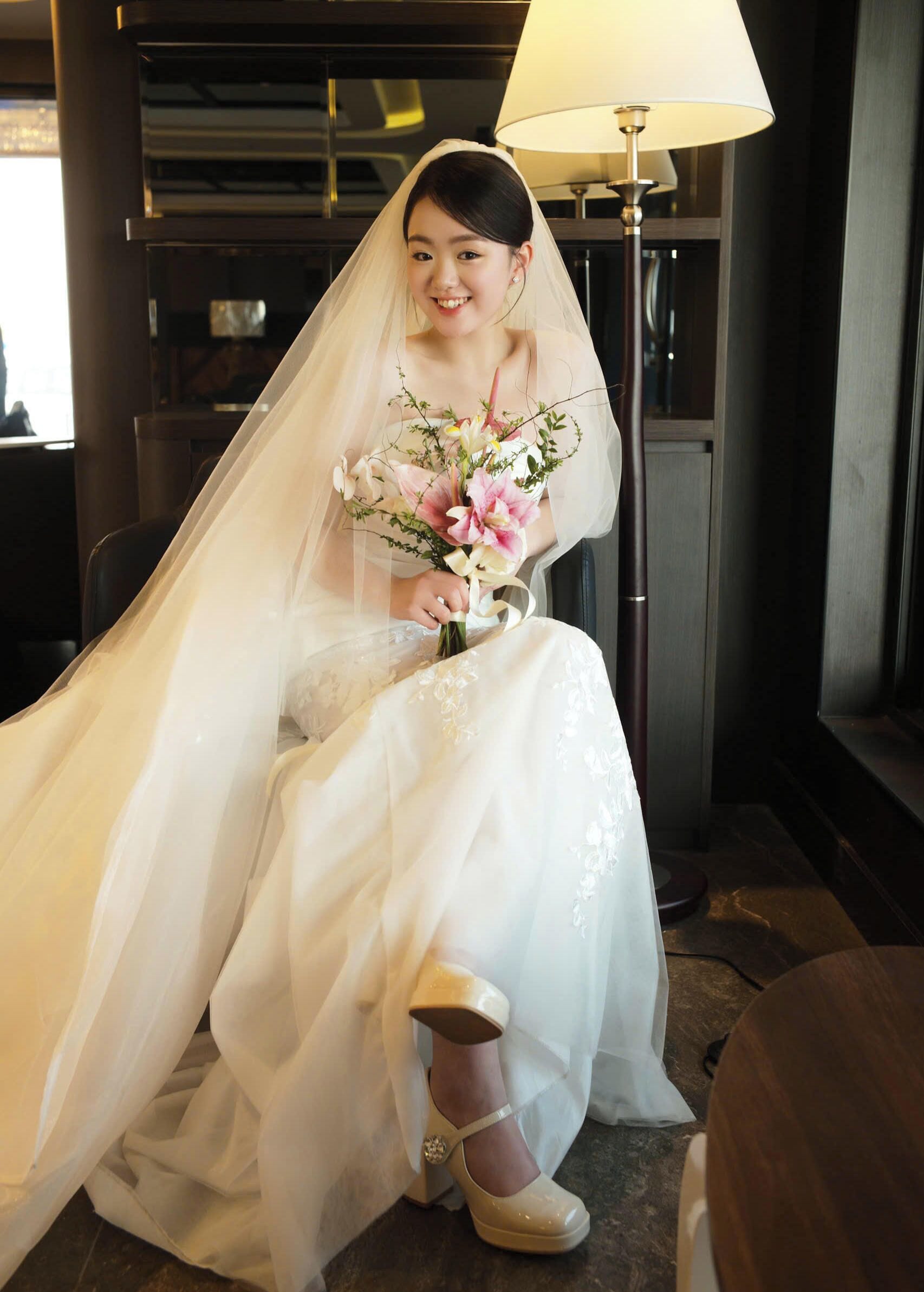 A person in a white wedding dress with a veil, holding a bouquet, seated in a room with a lamp and a wooden floor.