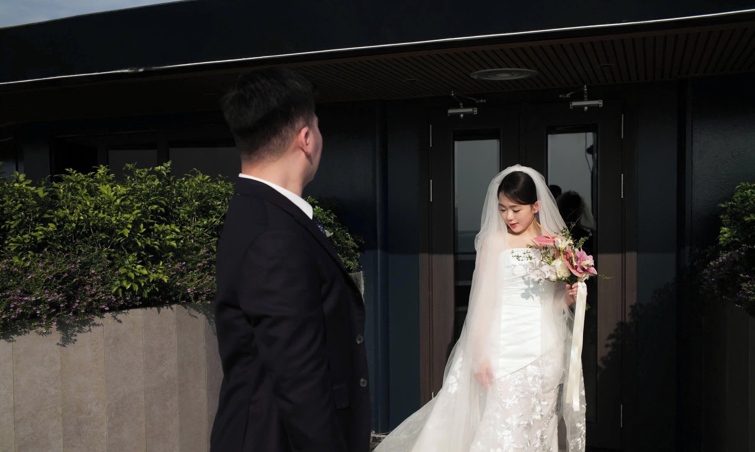 A couple in wedding attire outside a building with a modern design, greenery, and a clear sky.