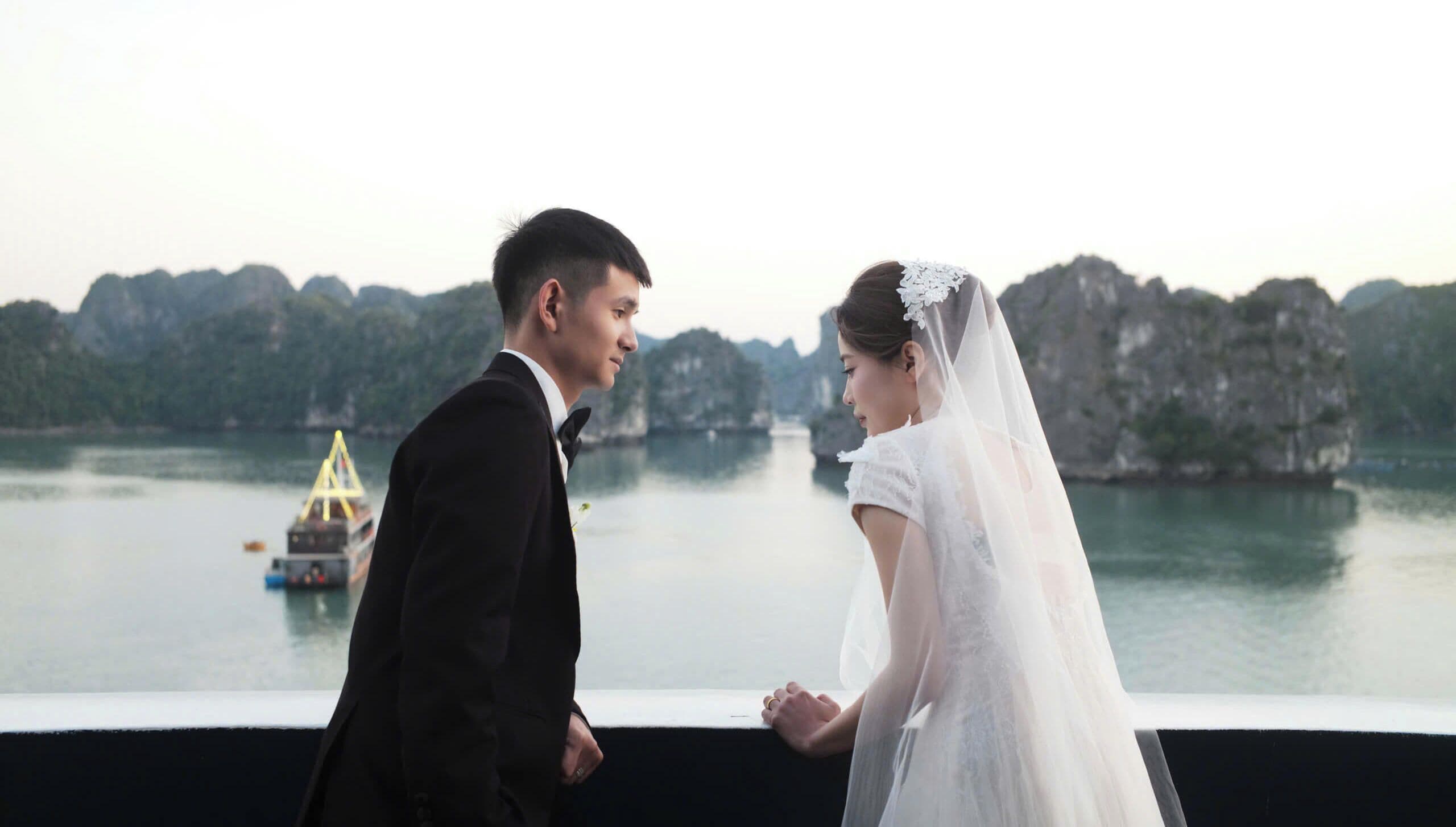 A couple holding hands by a waterfront with rocky cliffs in the background.