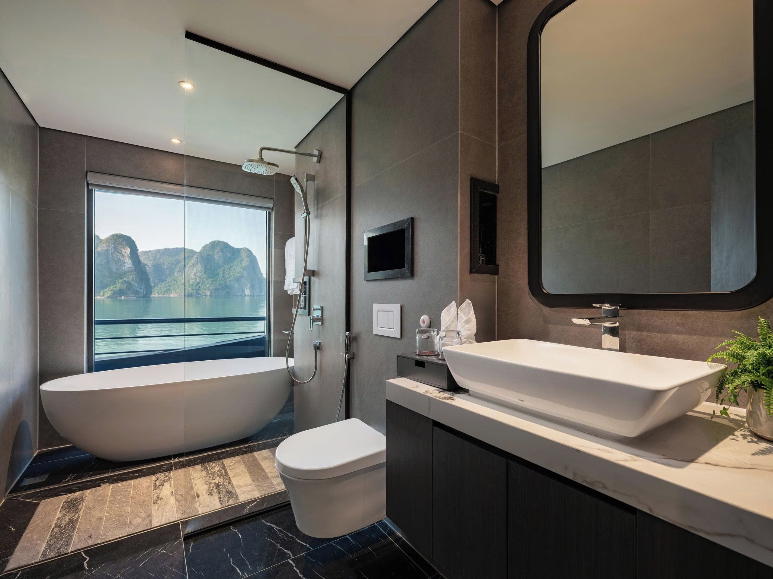 Un baño moderno con bañera, inodoro, lavabo y un espejo grande, con vistas a un paisaje montañoso a través de un ventanal.