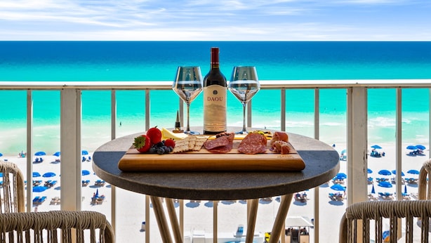 A table with a bottle of wine, two glasses, and an assortment of cheeses and meats on a balcony overlooking a beach.