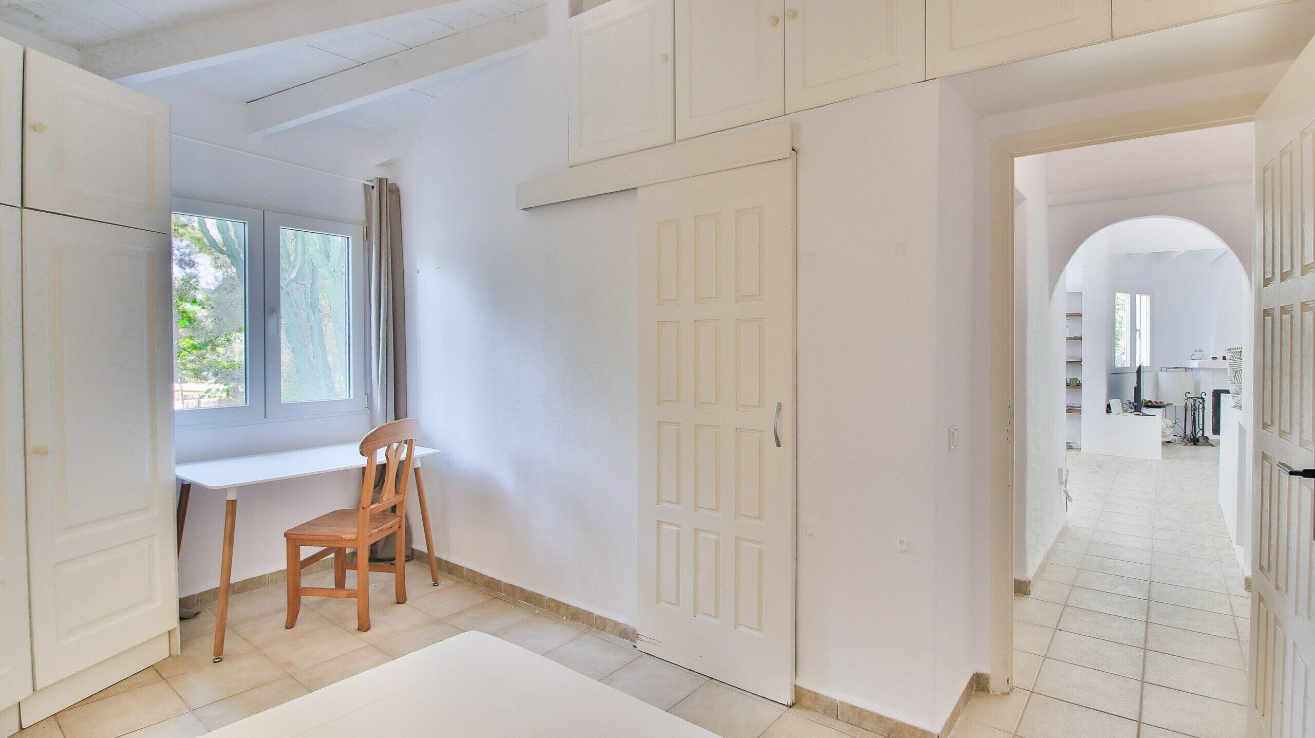 A room with a desk, chair, and wardrobe. There is a window with curtains, and a door leading to another room.