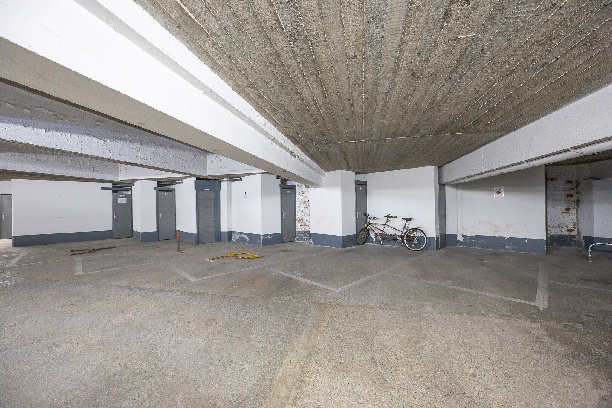 Un parking souterrain vide, avec des plafonds et des murs en béton, un vélo et un outil jaune posé sur le sol.