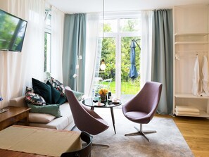 Een moderne woonkamer met een bank, twee fauteuils, een salontafel, een televisie en uitzicht op een tuin door grote ramen.