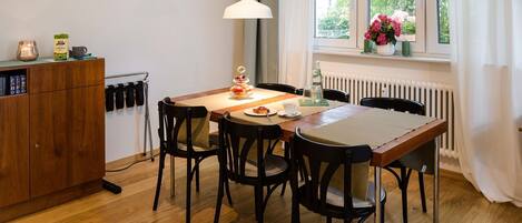 A dining area with a table, chairs, a lamp, a radiator, and a window with a view of greenery.