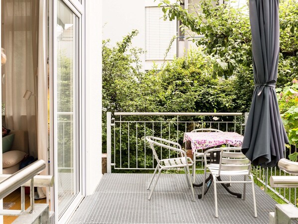 Een balkon met een metalen hekwerk, twee witte stoelen, een klein tafeltje met een tafelkleed met bloemenprint en een grijze parasol.
