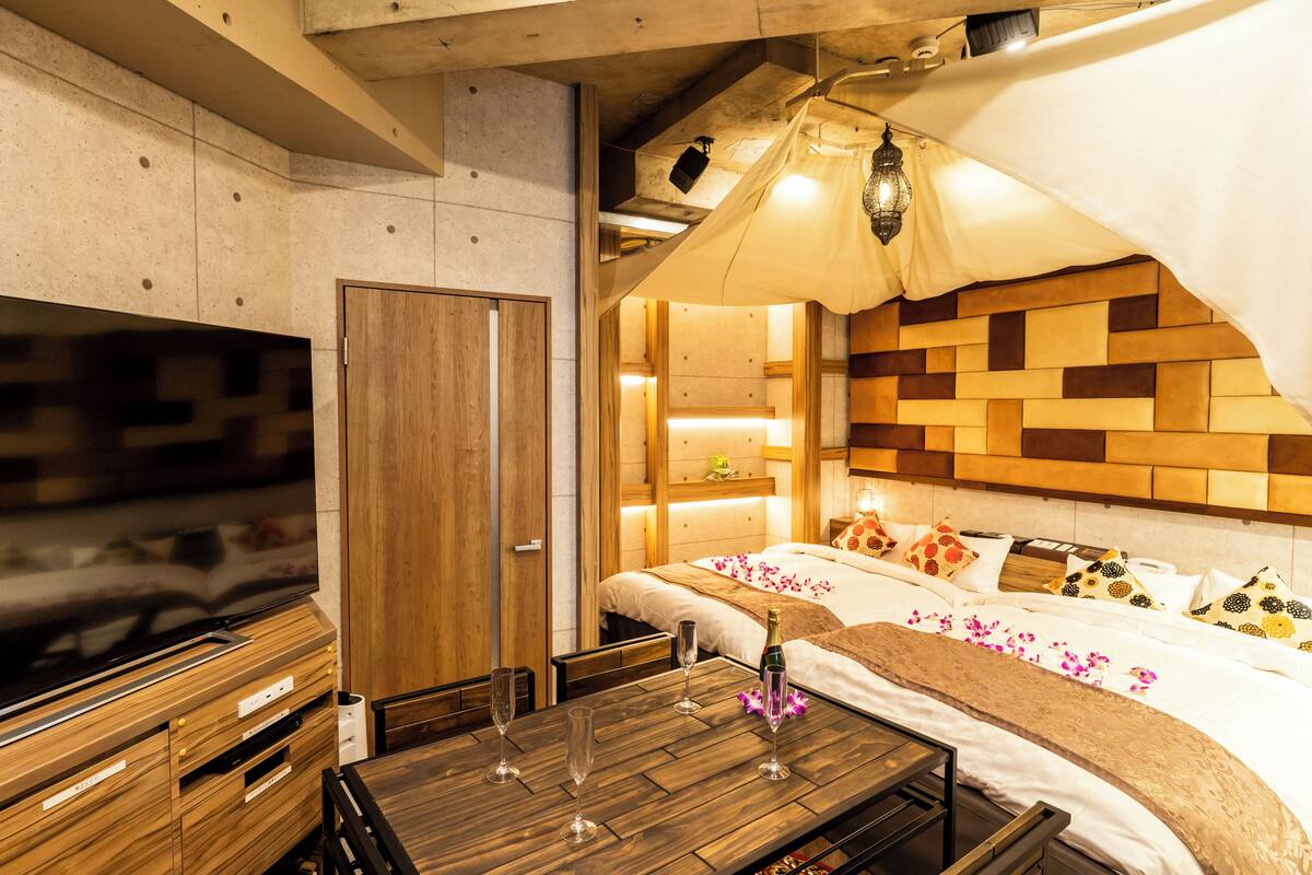 A modern hotel room with a wooden headboard, a flat-screen TV, a dining table with wine glasses, and a canopy bed.
