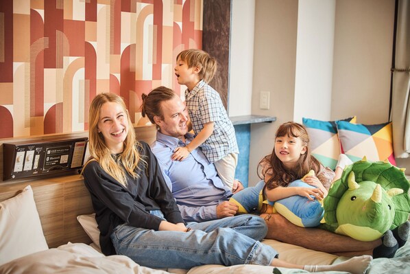 A family of four on a bed with a modern geometric wall design, a flat-screen TV, and a dinosaur plush toy. - Hotel BaliAn Resort - Shinjuku Forest (Tokyo)