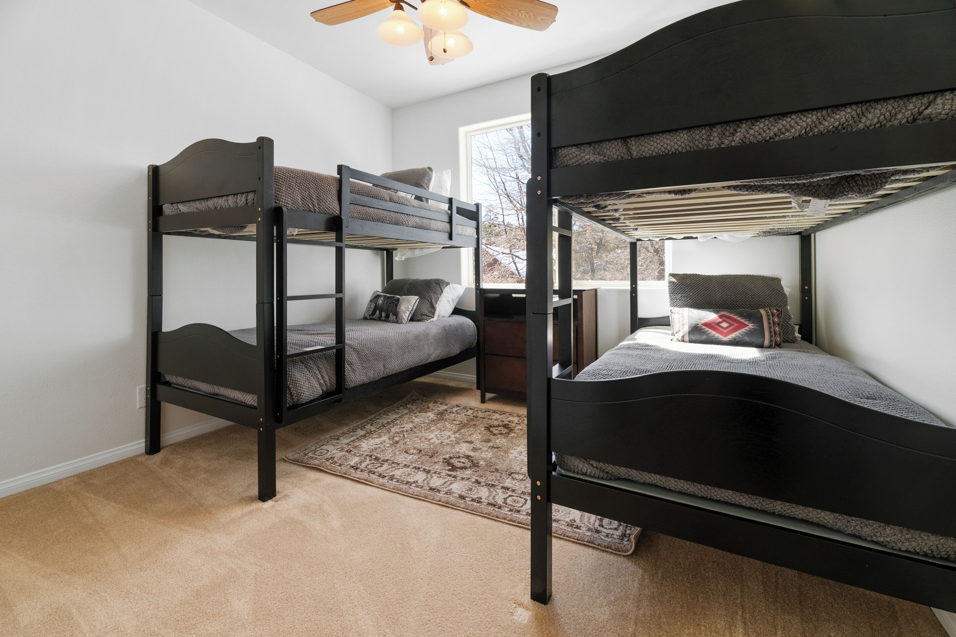 A bunk bed room with a ceiling fan, a window with a view, and a rug on the floor.