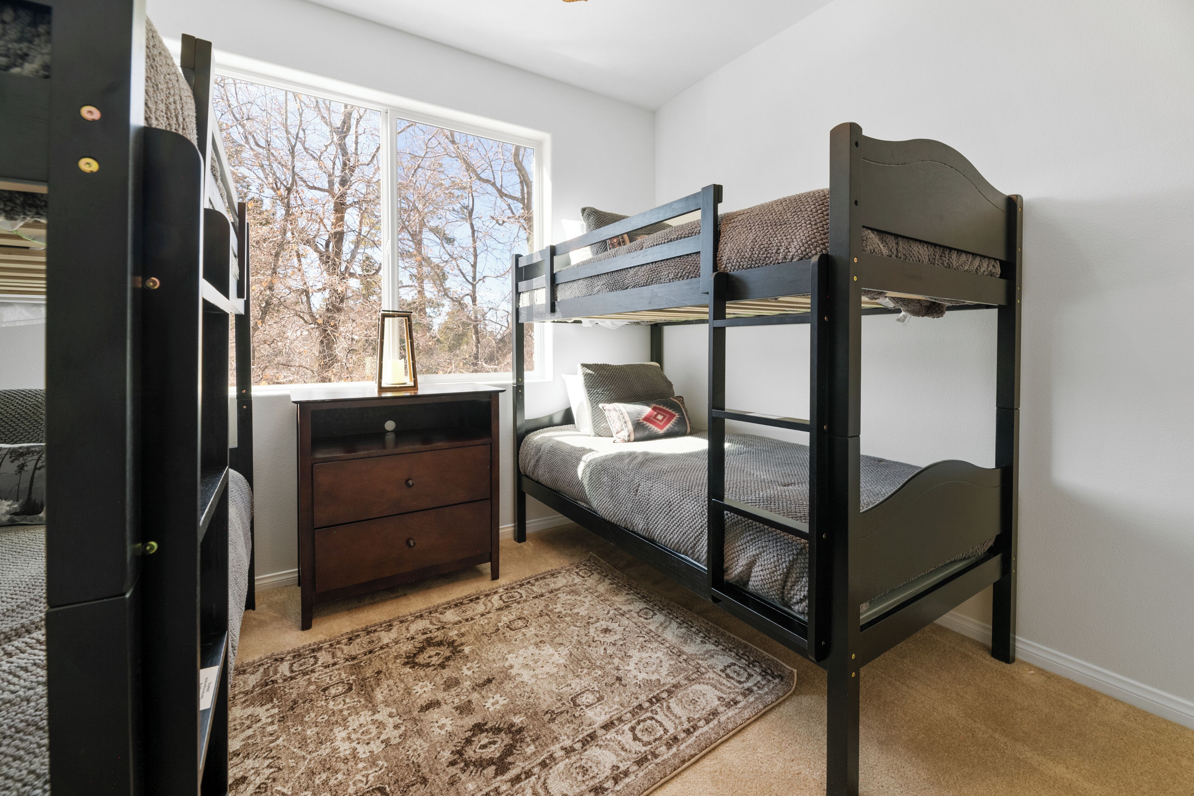 Een slaapkamer met een stapelbed, een bureau met stoel, een dressoir en een raam met uitzicht op bomen.