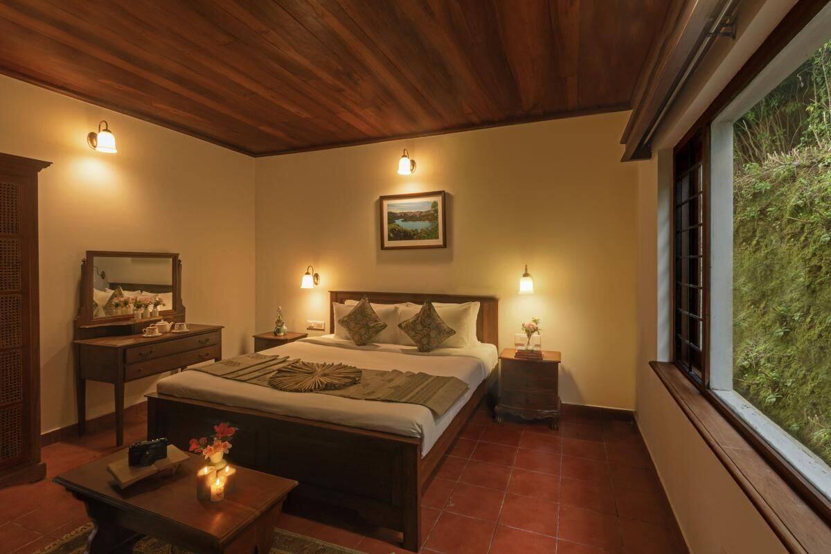 Un dormitorio con techo de madera, una cama grande con almohadas, mesitas de noche con lámparas, un aparador y una ventana con vista a zonas verdes.