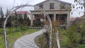 A paved pathway leading to a house with a garden and a cat walking along it.