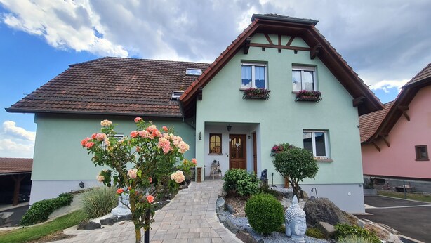 A two-story house with a tiled roof, a front entrance, and a garden with various plants and a Buddha statue.