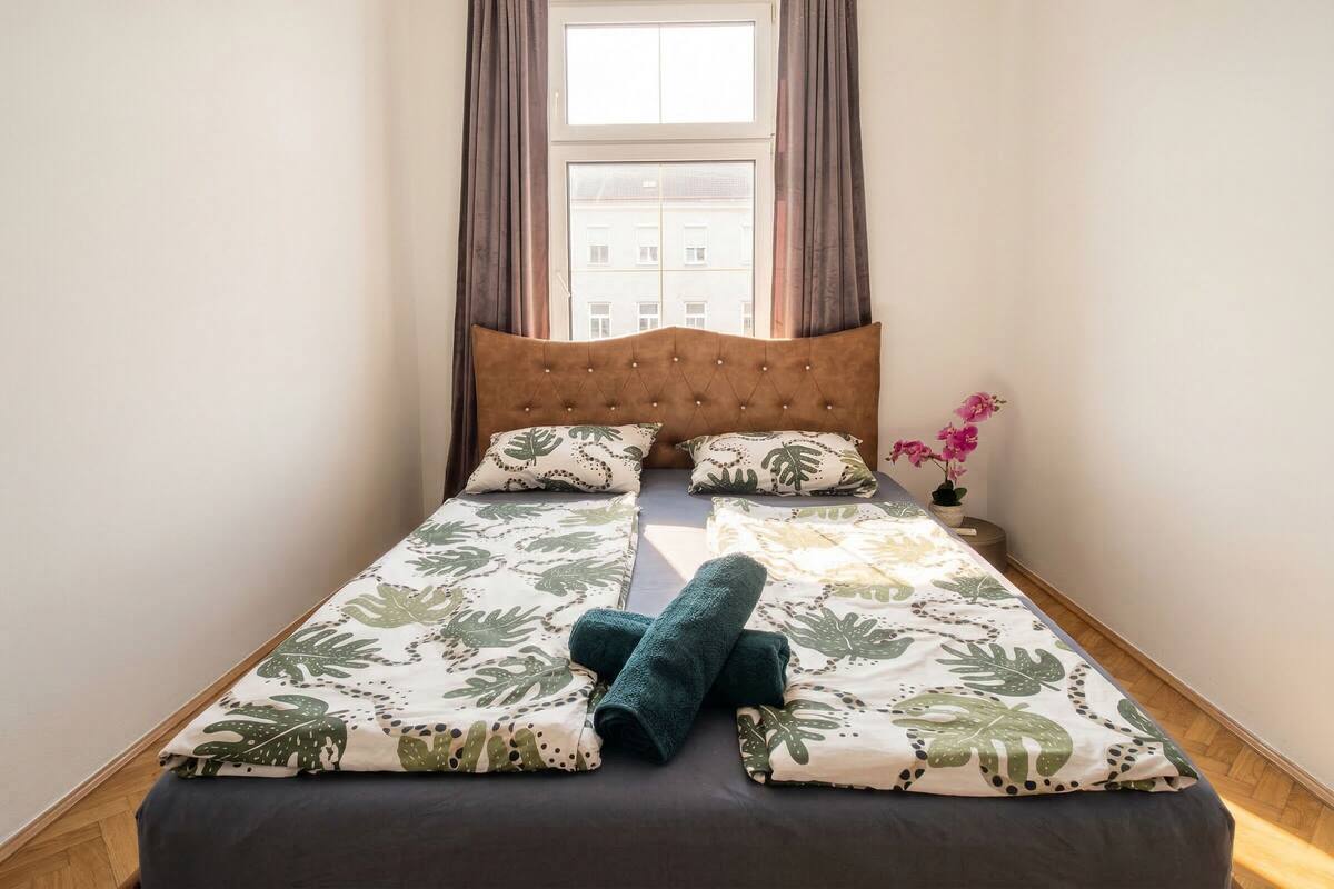 A neatly made bed with a leaf-patterned bedspread, a tufted headboard, and a window with curtains.