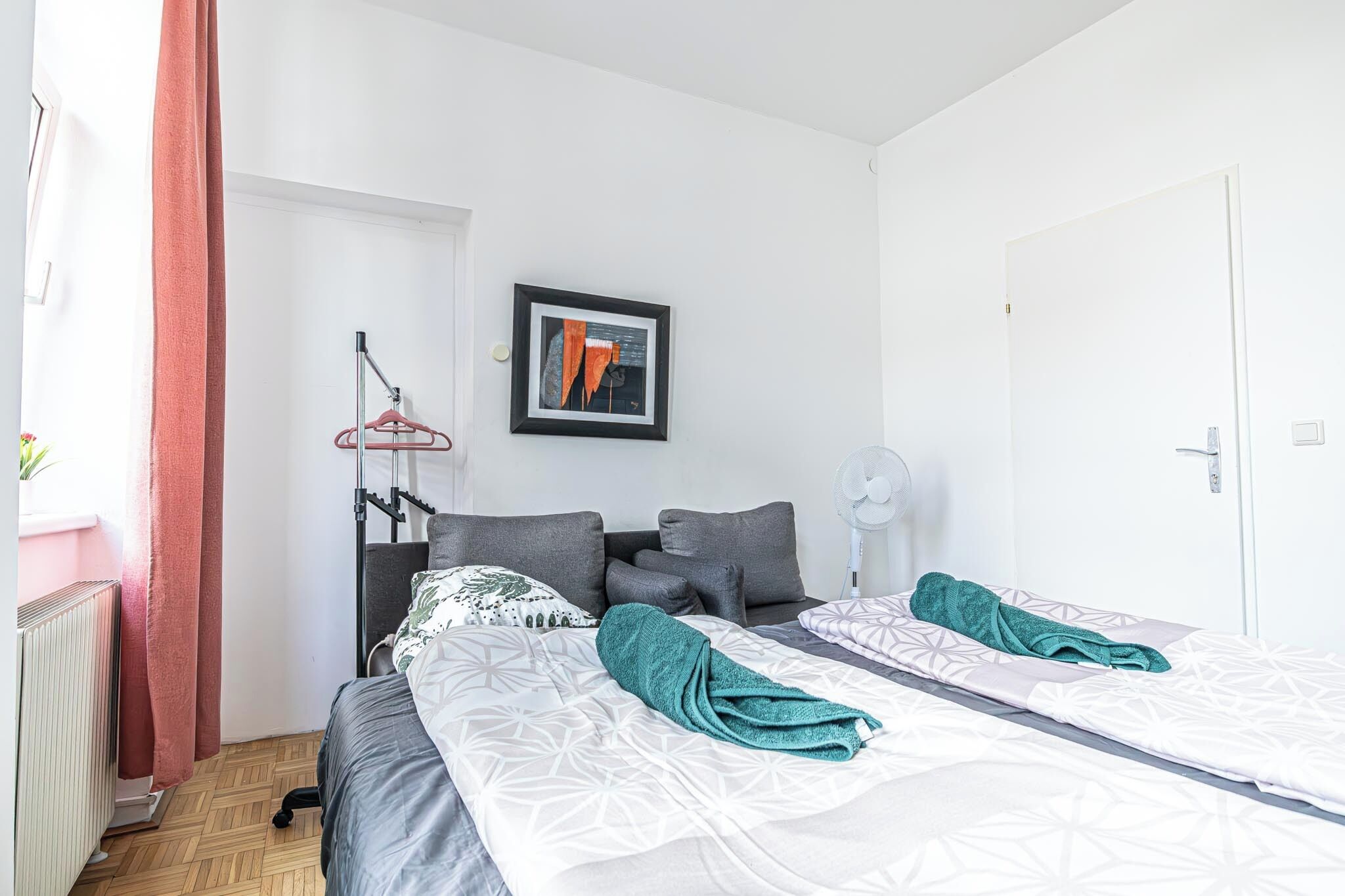 Una habitación de hotel moderna con una cama, un ventilador de pie, un radiador y un cuadro en la pared.