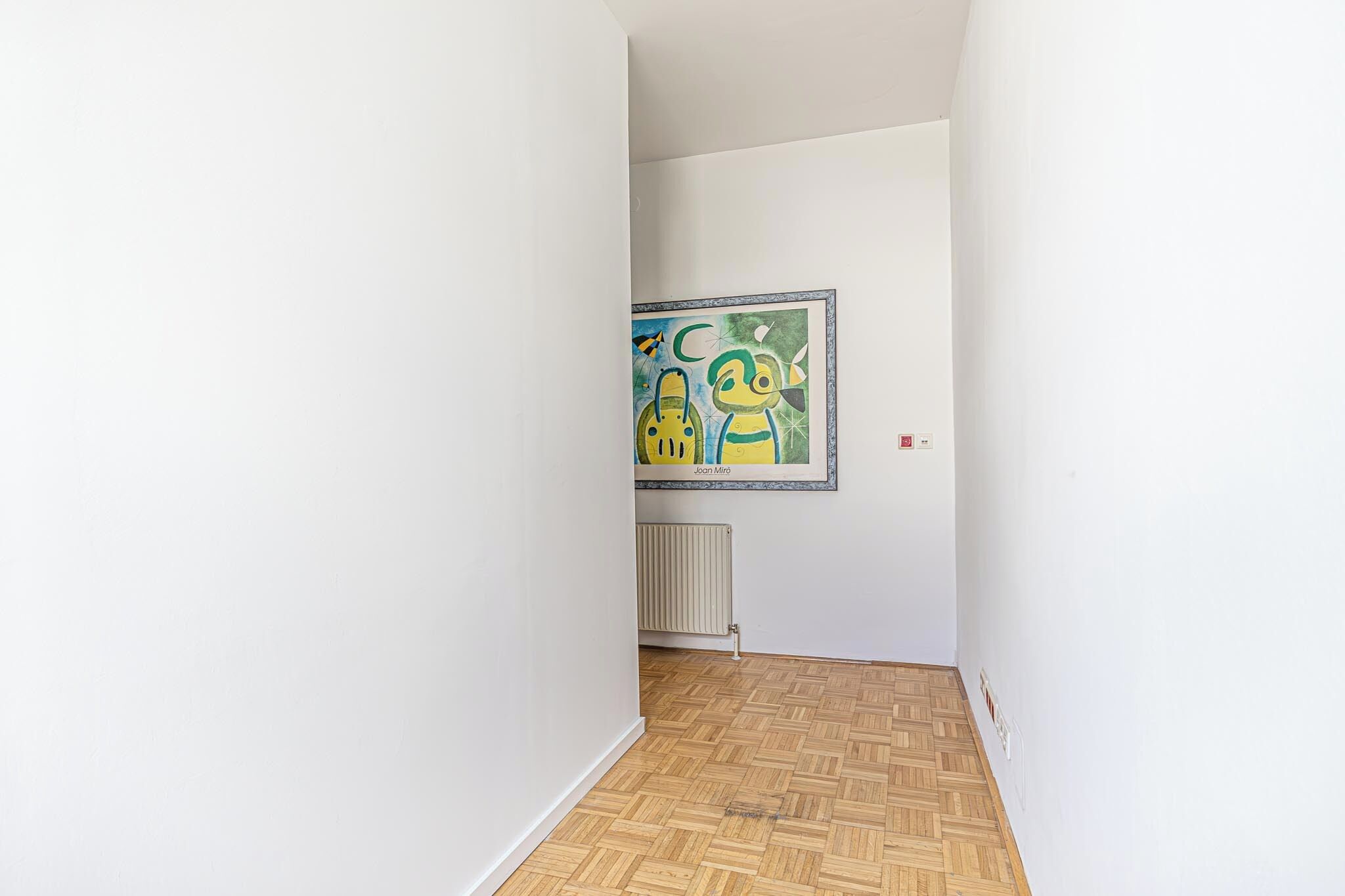 A hallway with a radiator, a framed artwork, and a herringbone-patterned floor.