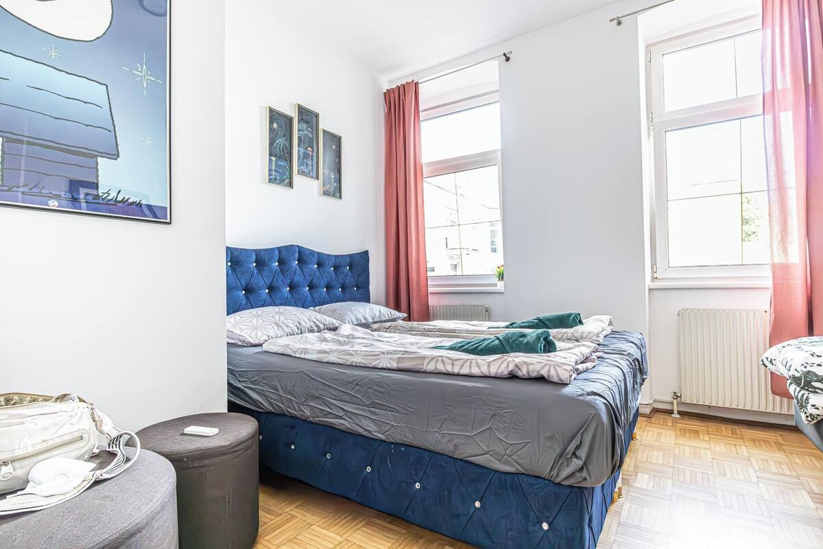 A bedroom with a blue headboard, a bed with grey and white bedding, a grey ottoman, a white bag, and a framed picture on the wall.