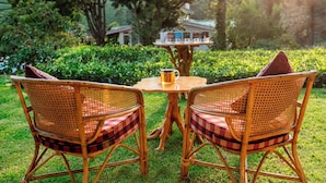 Two wicker chairs with checkered cushions and a wooden table with a mug on it, set in a lush garden with a small house in the background.