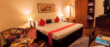 A hotel room with a bed, two bedside lamps, a wooden wardrobe, a small table with a chair, a bookshelf, and a framed picture on the wall.