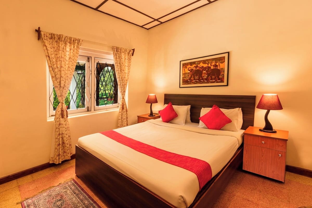 Une chambre à coucher bien rangée, avec un grand lit, deux tables de chevet avec des lampes, une fenêtre avec des rideaux et un tableau encadré au mur.